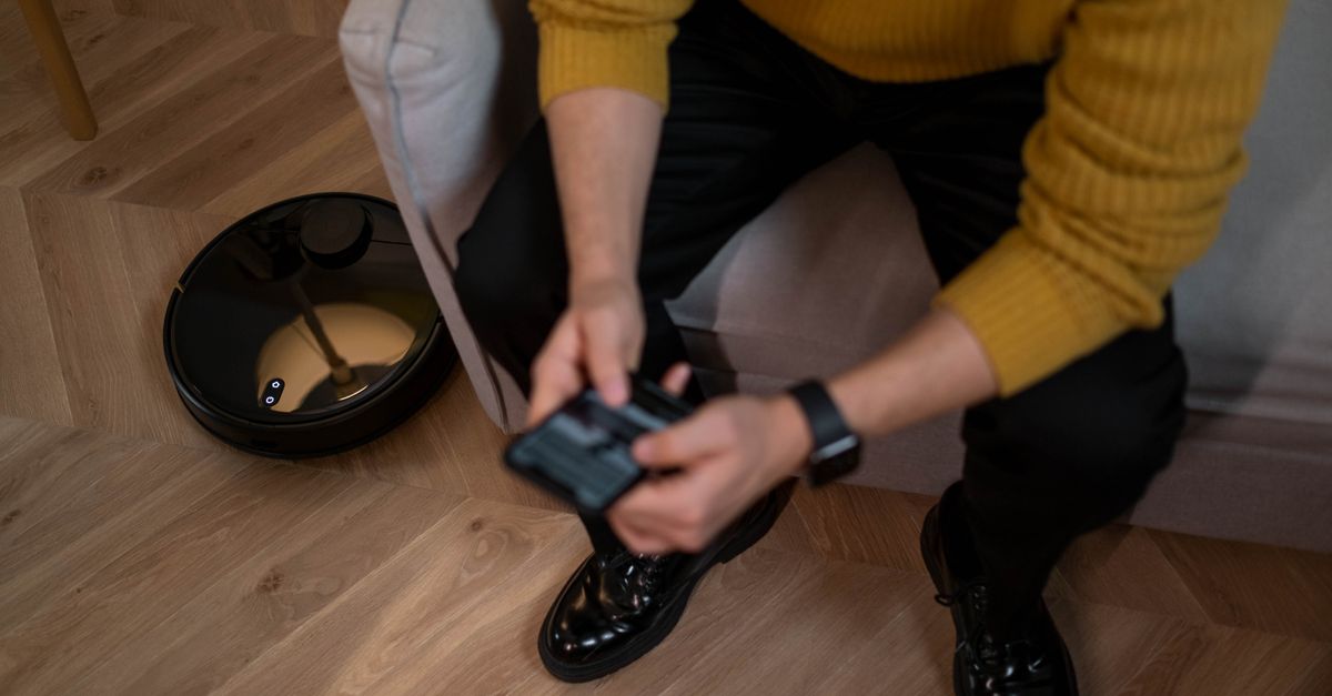Man in yellow sweater using remote to control robotic vacuum on wooden floor.